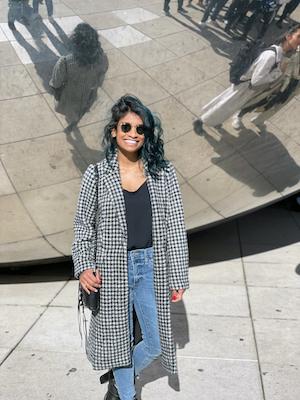 Deepa Lakshmin poses for a photo in front of a reflective surface, wearing jeans, a black top, a black-and-white coat, and a purse