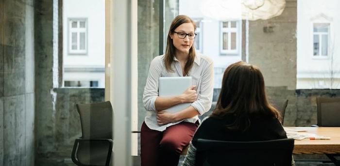 two people at an office talking, one sitting on a chair with their back to the camera and the other sitting on the table in front of them