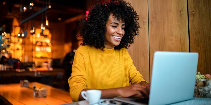 person sitting in a cafe smiling and typing on their laptop with a cup and saucer next to the computer