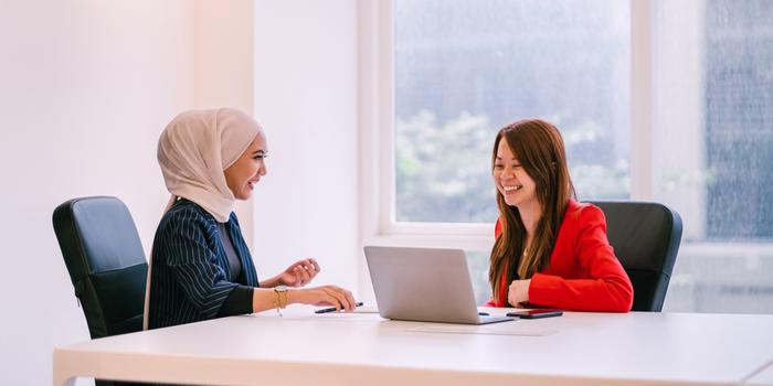 two people sitting at the corner of conference table laughing during job interview. laptop on table, windows in background