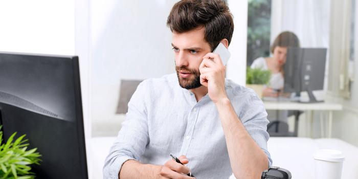 person sitting at desk, talking on the phone in front of a computer with another person visible in the background in another office