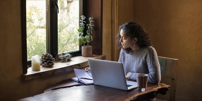 person sitting at table next to window, looking at notebook with laptop open in front of them