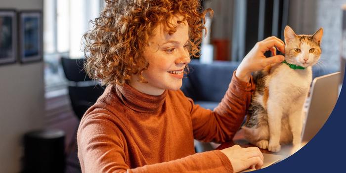 person sitting down in front of their open laptop while working from home, one hand on the keyboard, the other hand petting a cat that's sitting next to their laptop