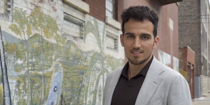 a person with curly hair wearing a light gray blazer standing in front of an alley