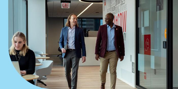 Two CDW employees walking down a hallway while engaged in conversation; on the far left, an employee sits at a table in front of a laptop.