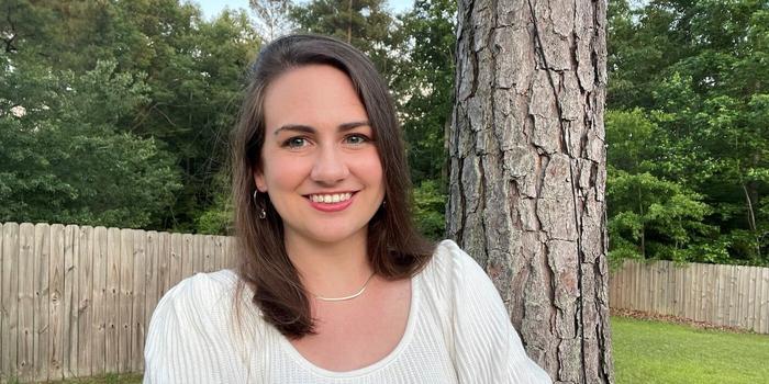 a person with shoulder-length brown hair, smiling in front of a tree in a backyard