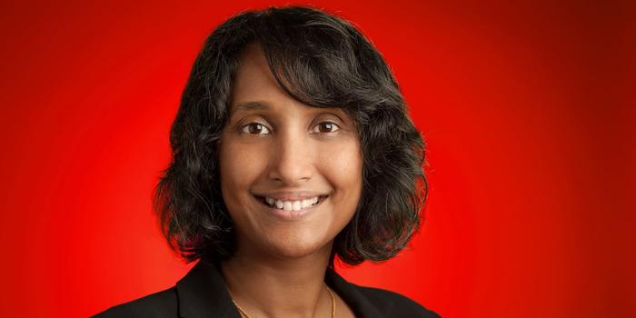 a person with dark wavy hair wearing a blazer and smiling in front of a red background