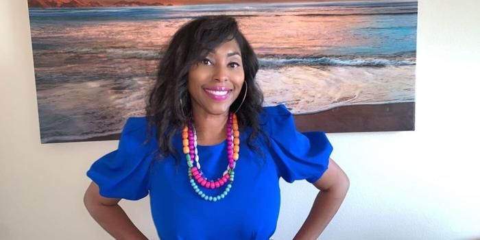 a person with a bight blue blouse and colorful necklaces smiling in front of a painting