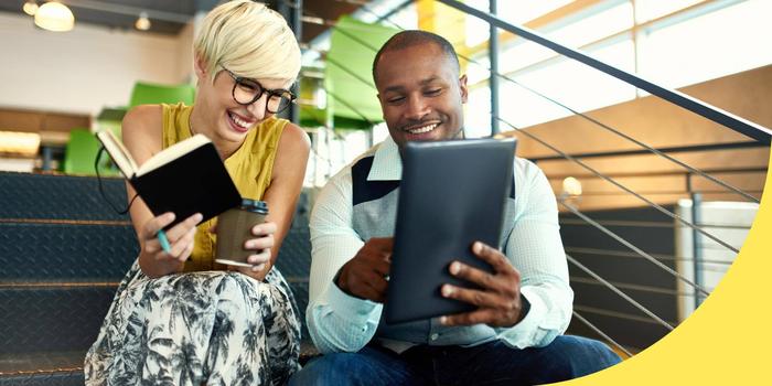 two smiling coworkers sitting on stairs in the office, one holding a tablet they're both looking at and the other holding a notebook and coffee cup