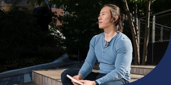 Jason Shen sits on steps outside, holding a notebook in his hands, looking off outside the frame, with trees in the background