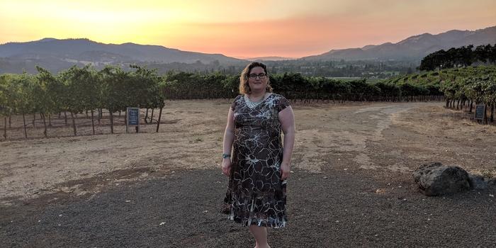 person smiling and standing in front of a vineyard during sunset