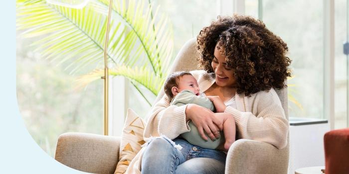 A woman with curly hair sitting in a chair at home, holding her baby.