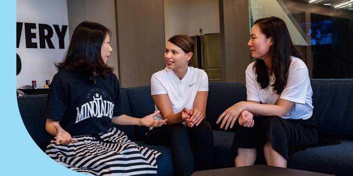Three people sitting on a couch and having an animated discussion. The person on the left is dressed in a T-shirt and a skirt; the person in the middle is dressed in a T-shirt and pants; the person on the right is wearing a T-shirt and a skirt.