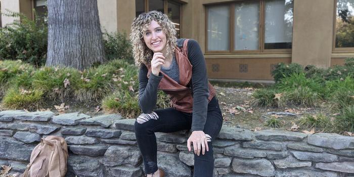 smiling person with curly hair sitting on a stone wall