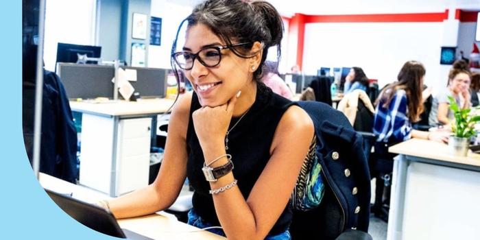 A photo of a smiling person sitting in front of an open laptop and computer monitor. The person is wearing glasses, a black tank top, and jeans.