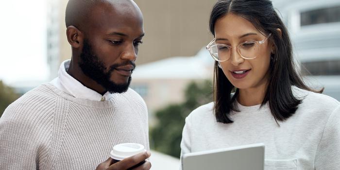 two coworkers standing outside on a balcony looking at a tablet; one of them is holding a cup of coffee