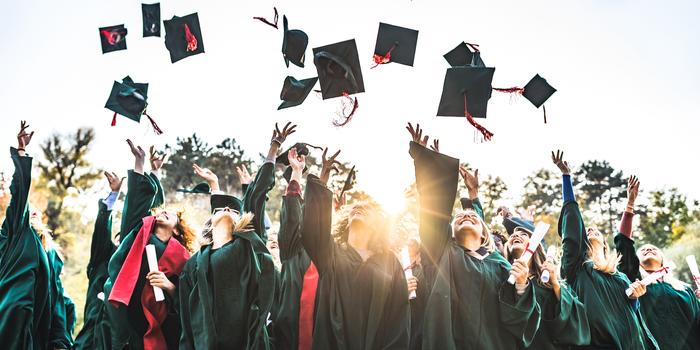 group of graduates in green robes throwing graduation caps in the air with treetops and bright sun in the background