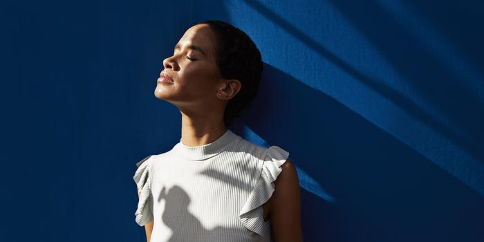 person with eyes closed standing against blue background and turned to the side, mostly in shadoww, with light from window on their face.