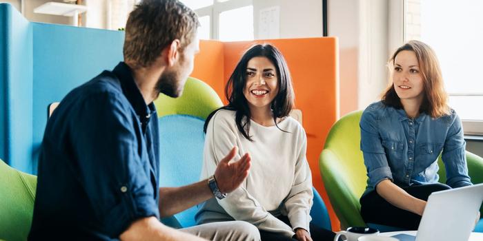 three colleagues sitting on brightly colored chairs in an office with an open laptop in front of them as they work on a cross-functional project