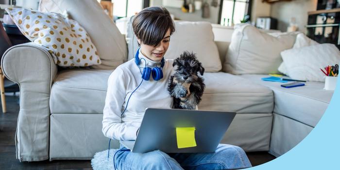 person sitting on the floor in front of a gray couch, looking at a laptop with a small dog in one arm