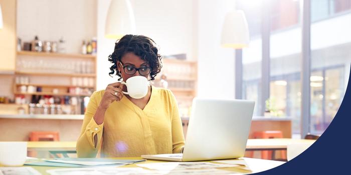 person sitting at a table in a light-filled room looking at an open laptop while sipping from a mug