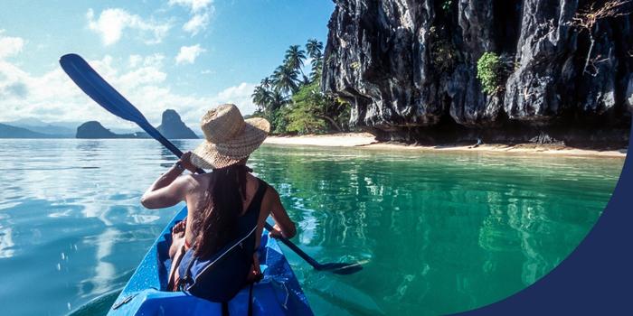 back of a person sitting in the front of a two-person kayak, paddling through blue-green water, with a large cliff face on the right and palm trees and mountains in the distance