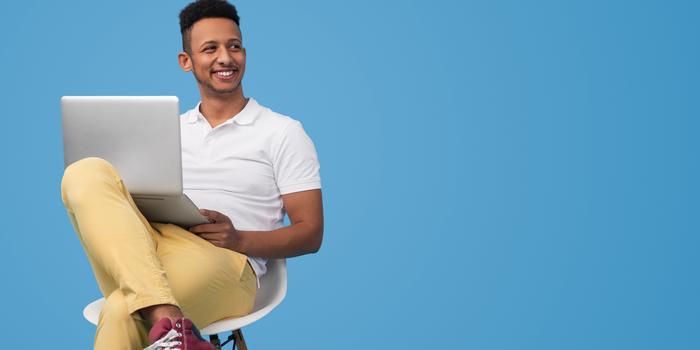 person sitting cross-legged in a white chair against a blue backdrop, smiling and holding a laptop open