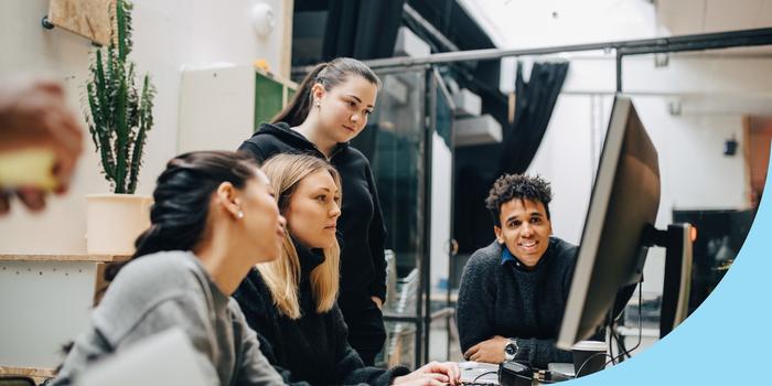 group of coworkers sitting and standing around a table in an office, looking at a computer screen