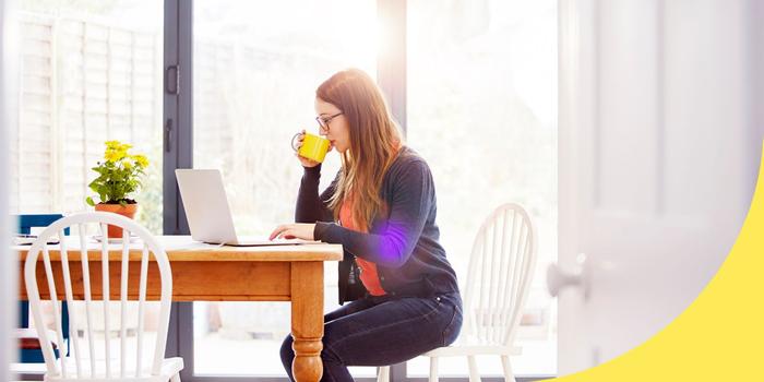 person sitting at their kitchen table, drinking from a mug, and working on a laptop