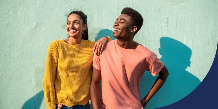 two happy young professionals standing next to each other, one in a yellow sweater, the other in a salmon-colored shirt, both smiling and looking off to the side against a green background