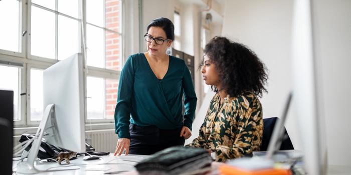 two coworkers looking at a computer in an office, one standing, one sitting