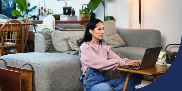 person sitting on the floor between a couch and a coffee table, typing on and looking at laptop