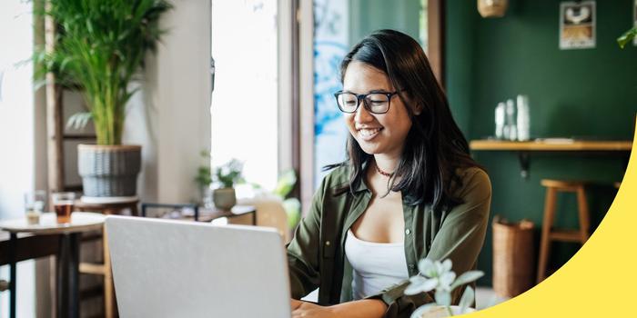 person sitting at table in coffee shop with dark green walls, typing on laptop and smiling