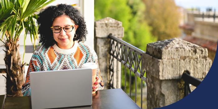 person working on a laptop while sitting at a table on a balcony with worn stone pillars and a plant in the background