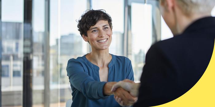 two people shaking hands in a job interview. One person is facing the camera with windows behind them.