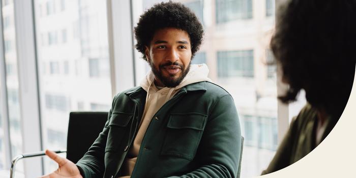 two people having a conversation in a conference room with large windows and buildings in the background; one person is facing the camera in a green jacket