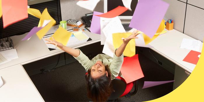 person sitting in an office cubicle, leaning back in their chair and smiling as they throw a stack of yellow, purple, red, and white paper in the air above them