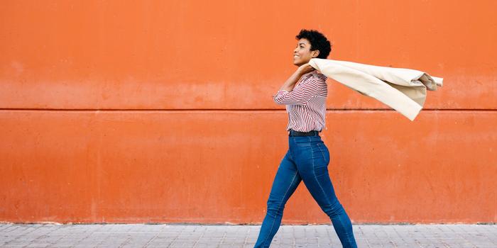 person walking down sidewalk in front of orange wall, holding a tan jacket over one shoulder