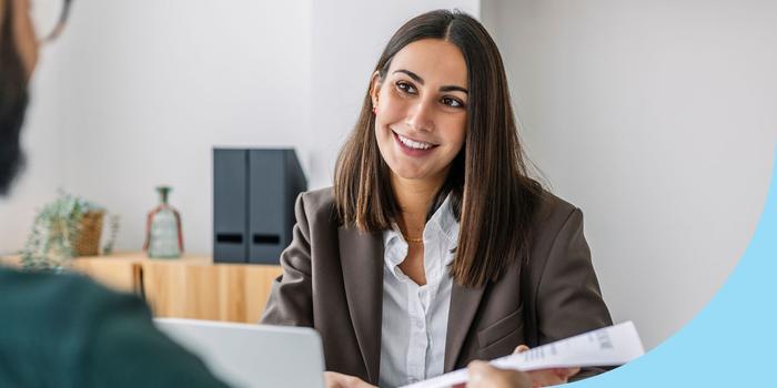 job applicant passing their resume to the hiring manager in a job interview in an office with white walls and a small wood shelf in the background