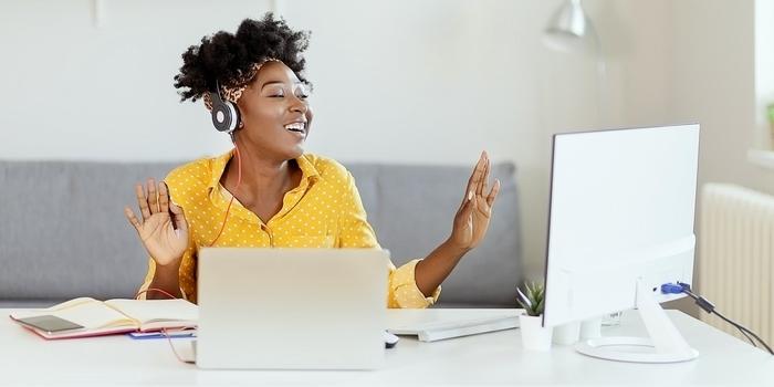 person wearing headphones, holding hands up slightly and singing, while sitting at desk with two computer monitors