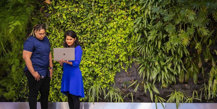 two people standing in front of a plant-covered wall, looking at a laptop together