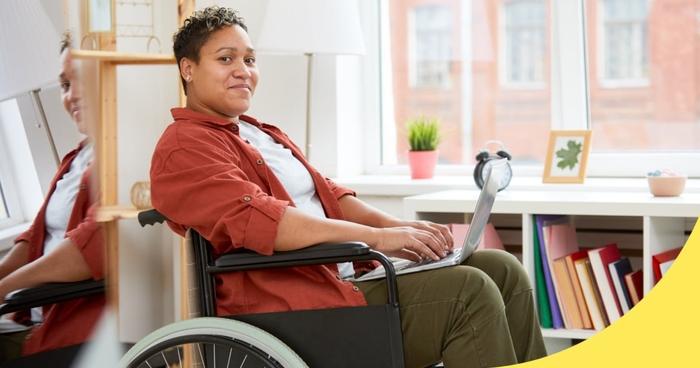 person sitting in a wheelchair with a laptop open on their lap, typing, with their office in the background
