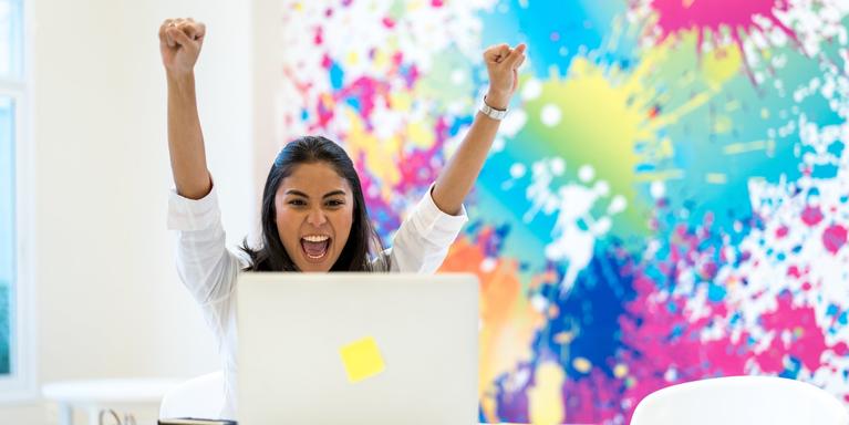 excited recruiter reading off a laptop screen with both arms in the air, multicolored splashes of paint in the background
