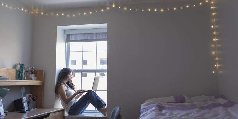 person sitting on windowsill in dorm room working on laptop