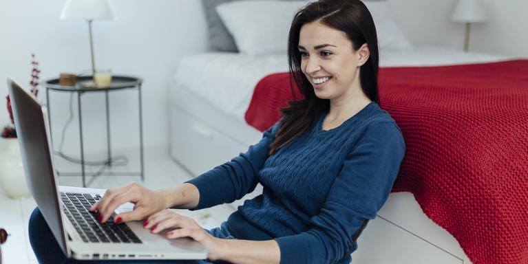 smiling person sitting on floor next to bed on laptop