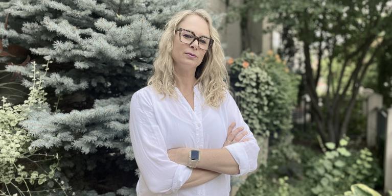 person with long blonde hair and black-framed glasses standing in front of trees