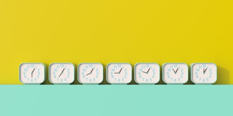 line of teal analog clocks set to different times on a teal surface against a yellow wall