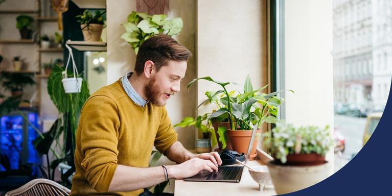 person sitting at a window table in a cafe typing on laptop, with two plants on the table and more in the background