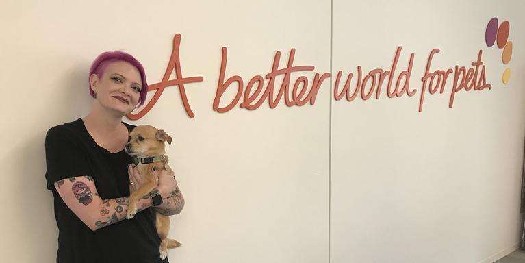 person with short pink hair holding a dog and standing against a wall that says "A better world for pets."