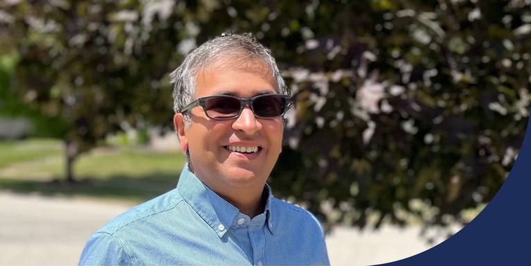 photo of Sameer Doshi wearing a blue button-up shirt and sunglasses and standing outside with trees and grass in the background